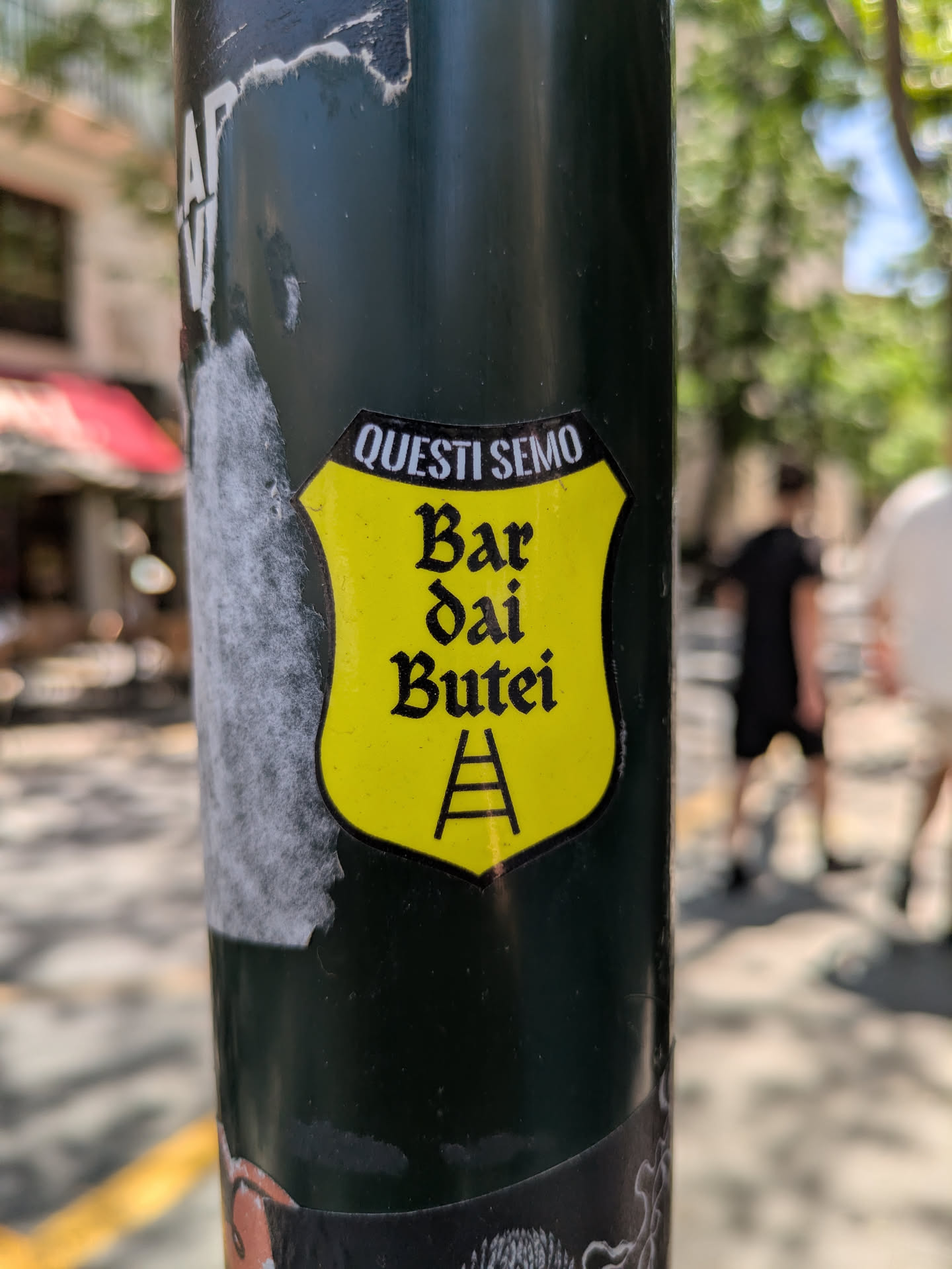 The sticker is on a metal pole. The sticker is yellow and shaped like a shield. The text "QUESTI SEMO" is at the top. Below that is "Bar dai Butei" and at the bottom is a drawing of a ladder.