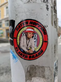 Street sticker The sticker is circular, predominantly red and yellow, featuring a photographic image of an Asian man wearing a red and white striped bucket hat. He appears pensive or serious. The outer red ring contains black text: 'HATER HOPPING' at the top and 'ELSKER BRANN' at the bottom. The inner yellow ring, which surrounds the man's image, has smaller black text that reads 'CIVITATE:BERGENS:SIGILLVM:COMMVN' along with a stylized depiction of a castle or city gate, commonly associated with the Bergen city seal. The abbreviation 'SPV' is also present on the red ring on either side of the man. The sticker is adhered to a weathered pole, with remnants of other stickers visible around it.