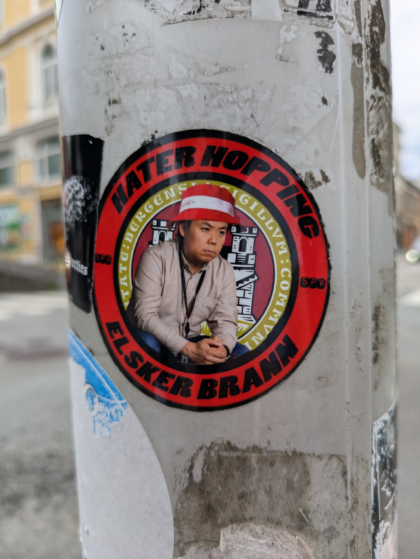 The sticker is circular, predominantly red and yellow, featuring a photographic image of an Asian man wearing a red and white striped bucket hat. He appears pensive or serious. The outer red ring contains black text: 'HATER HOPPING' at the top and 'ELSKER BRANN' at the bottom. The inner yellow ring, which surrounds the man's image, has smaller black text that reads 'CIVITATE:BERGENS:SIGILLVM:COMMVN' along with a stylized depiction of a castle or city gate, commonly associated with the Bergen city seal. The abbreviation 'SPV' is also present on the red ring on either side of the man. The sticker is adhered to a weathered pole, with remnants of other stickers visible around it.