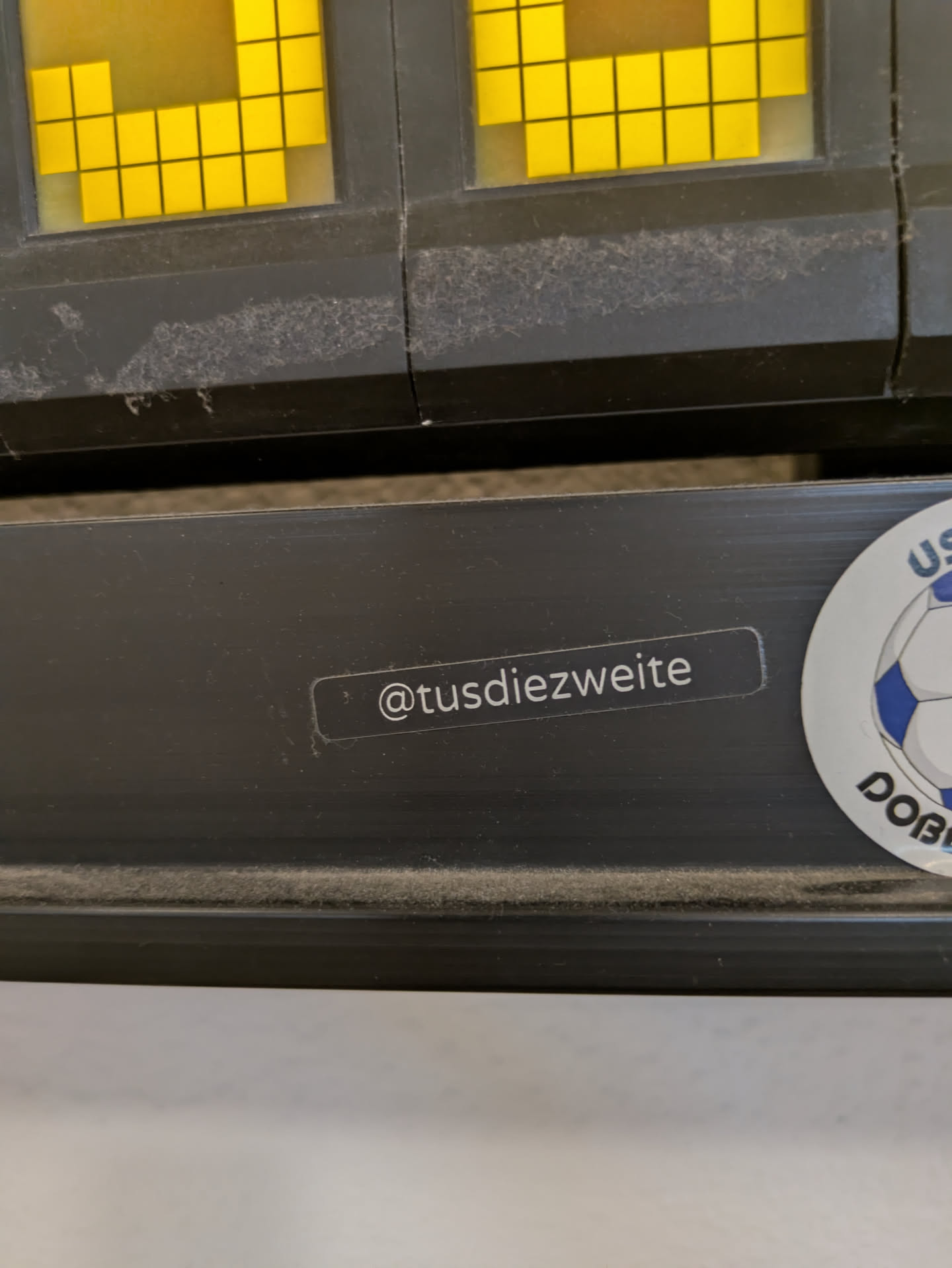 The image features two stickers on a dark surface. One sticker contains text that reads '@tusdiezweite'. The other one is a sports logo. The background appears to be part of a scoreboard or similar display.