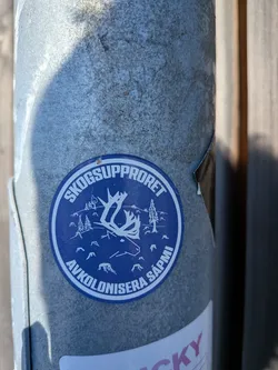Street sticker The sticker is circular with a dark blue background and white graphics. The central image depicts a stylized reindeer head with prominent antlers. Surrounding the reindeer are silhouetted pine trees and smaller representations of stumps, suggesting a forest environment. The text 'SKOGSUPPRORET' is written in a white arc at the top of the circle, and 'AVKOLONISERA SÁPMI' is written in a similar arc at the bottom. The overall design is graphic and impactful, with a clear environmental and political message.