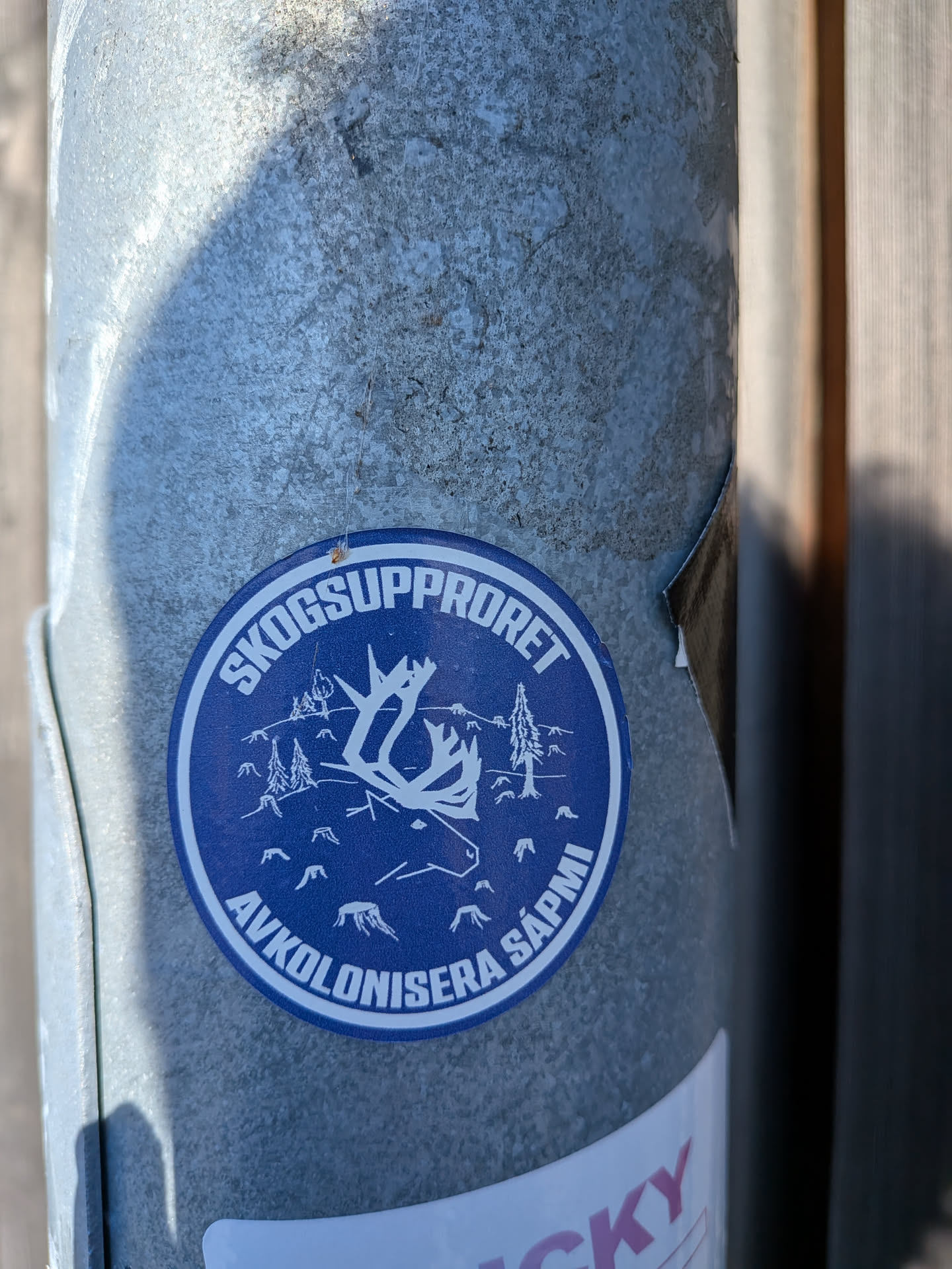 The sticker is circular with a dark blue background and white graphics. The central image depicts a stylized reindeer head with prominent antlers. Surrounding the reindeer are silhouetted pine trees and smaller representations of stumps, suggesting a forest environment. The text 'SKOGSUPPRORET' is written in a white arc at the top of the circle, and 'AVKOLONISERA SÁPMI' is written in a similar arc at the bottom. The overall design is graphic and impactful, with a clear environmental and political message.