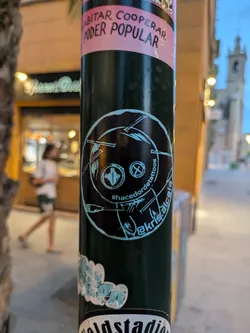 This round sticker features a dark background with a light blue artistic drawing resembling a face. The art style is somewhat primitive and minimalistic, featuring simple lines and shapes. Text along the border says '#hacedordetattoos' and '@akris.alcala'. It is affixed to a dark street pole among other stickers.