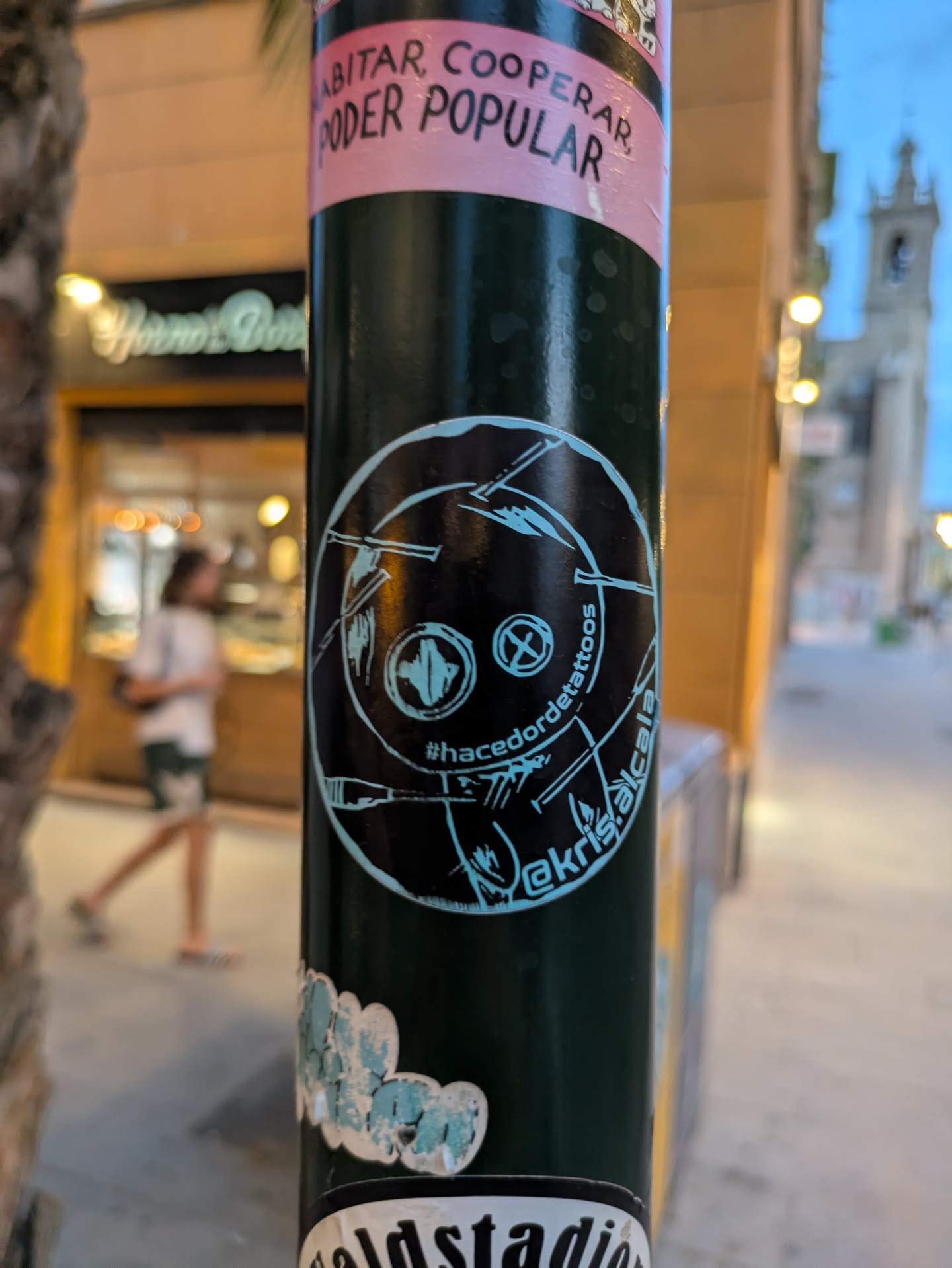 Street sticker This round sticker features a dark background with a light blue artistic drawing resembling a face. The art style is somewhat primitive and minimalistic, featuring simple lines and shapes. Text along the border says '#hacedordetattoos' and '@akris.alcala'. It is affixed to a dark street pole among other stickers.
