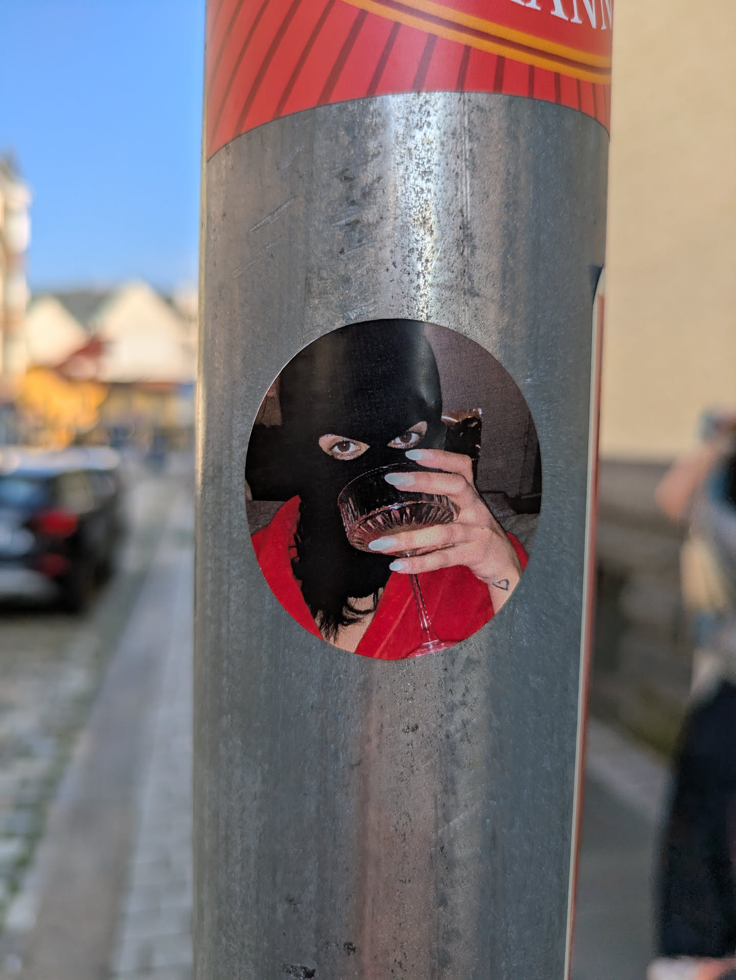 The sticker, which is circular and affixed to a metal pole, features a person wearing a black balaclava, with only their eyes visible. The individual is holding a glass filled with a dark liquid, possibly red wine, in their left hand. Their nails are long and painted a light, almost translucent, color. A small heart tattoo is visible on their inner wrist. The person is wearing a red top, contrasting with the black balaclava. The image evokes a sense of anonymity and intrigue.