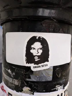 Street sticker The sticker features a high-contrast, black and white stylized portrait of a person's face, likely a woman, with long, dark, flowing hair. The face is rendered with bold, graphic lines, reminiscent of a woodcut or stencil. The eyes are particularly striking, appearing wide and intense with white pupils against dark irises, conveying a piercing gaze. The mouth is slightly open, revealing a hint of white. The sticker itself is die-cut around the shape of the head, with a thin white border, and is placed on a larger, rectangular white background sticker. Below the chin, the text '@MIHAN.TATTOO' is visible in black.