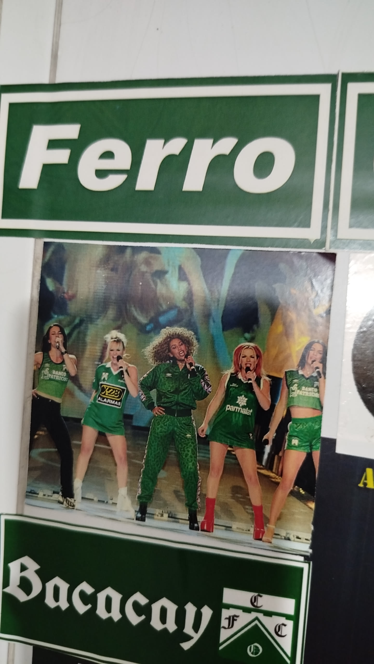 The central sticker features a vibrant photograph of the five members of the Spice Girls performing on stage. All five women are holding microphones and are dressed predominantly in green outfits, some with white or black accents. From left to right, the first woman wears a green tank top and black pants, the second a green mini-dress, the third a green tracksuit, the fourth a green mini-dress, and the fifth a green crop top and shorts. The stage background is a blurry mix of blues, yellows, and oranges, suggesting dynamic lighting. The image captures a moment of energetic performance.