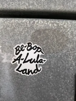 Street sticker A white sticker with black text that says \"Be-Bop-A-Lula-Land\" in a handwritten style. The sticker is affixed to a gray metallic surface.