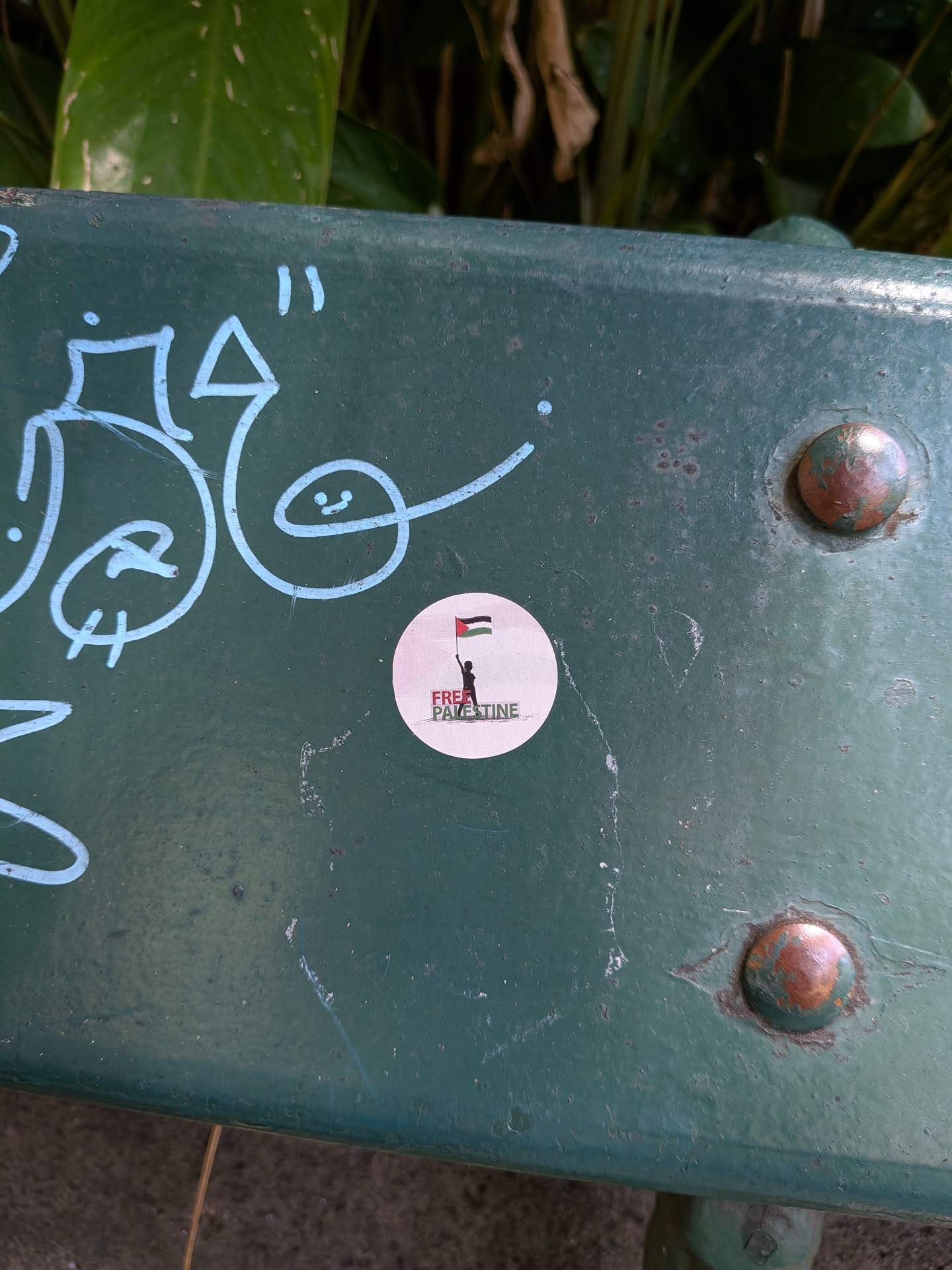 A circular sticker on a green metal bench. It depicts a black silhouette holding a Palestinian flag above the text 'FREE PALESTINE' in red and green.