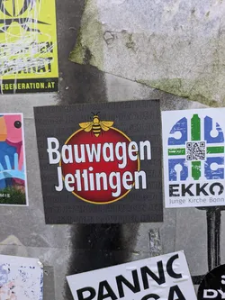 Street sticker A square, dark grey sticker centered on a wall, featuring a prominent red circle with a thin yellow outline. Inside the circle, the words "Bauwagen Jettingen" are written in bold white letters. A yellow bee graphic is positioned directly above the red circle. The dark grey background of the sticker is subtly patterned with faint, repeating text that also reads "BAUWAGEN JETTINGEN".