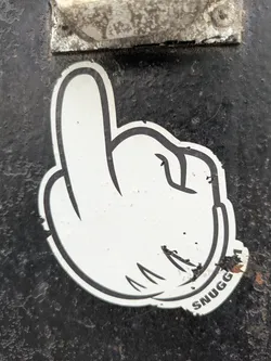 Street sticker This sticker features a white cartoon hand in a distinctive gesture, likely a play on a common rude hand sign, with black outlines. The word "SNUGG" is written in bold black letters at the bottom right corner of the sticker. The sticker appears slightly worn with some small damages.