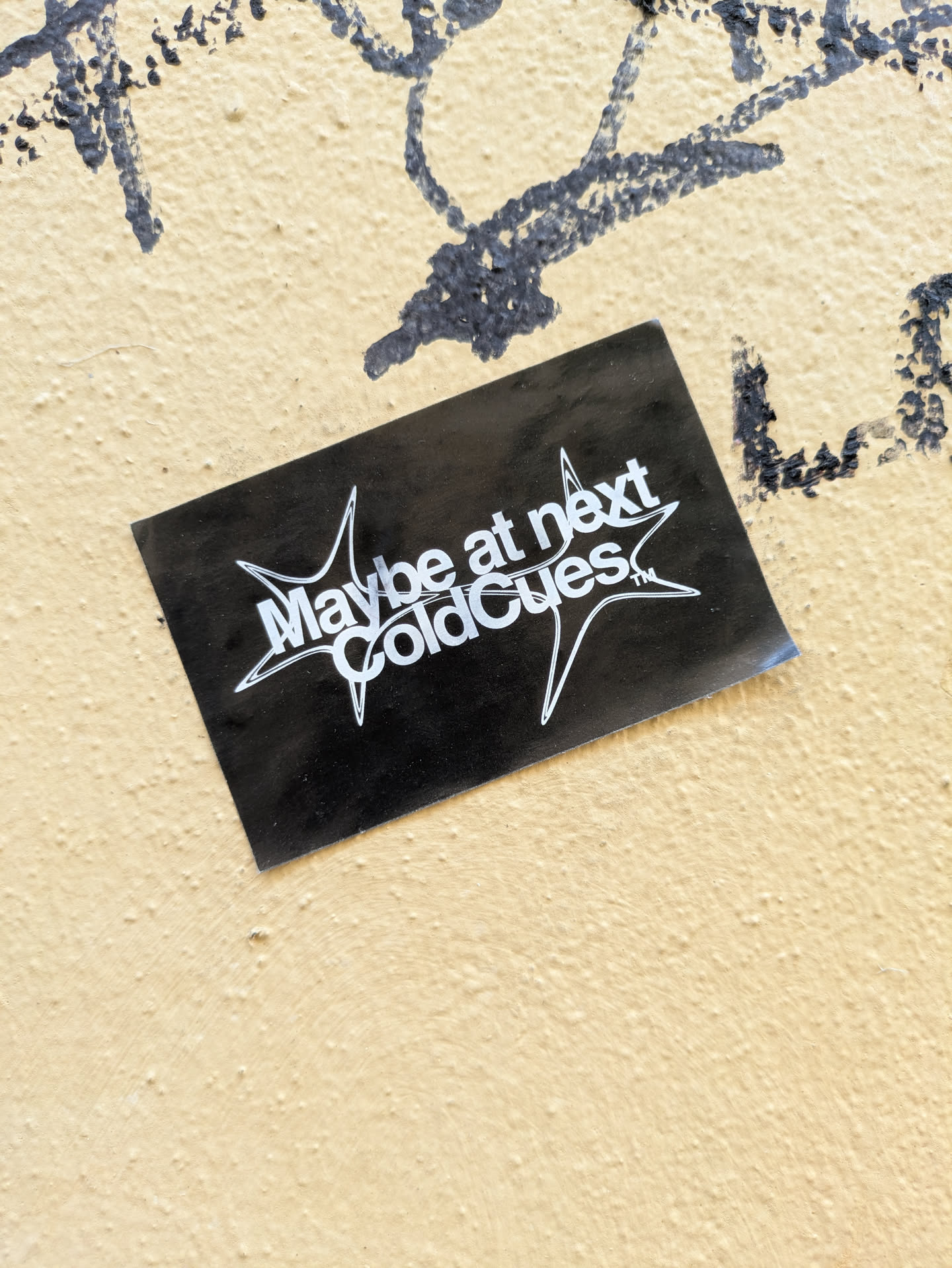 The sticker is black with white stylized text that reads 'Maybe at next ColdCues'. It appears to be placed on a yellow wall that has some black graffiti on it. The sticker is rectangle-shaped.