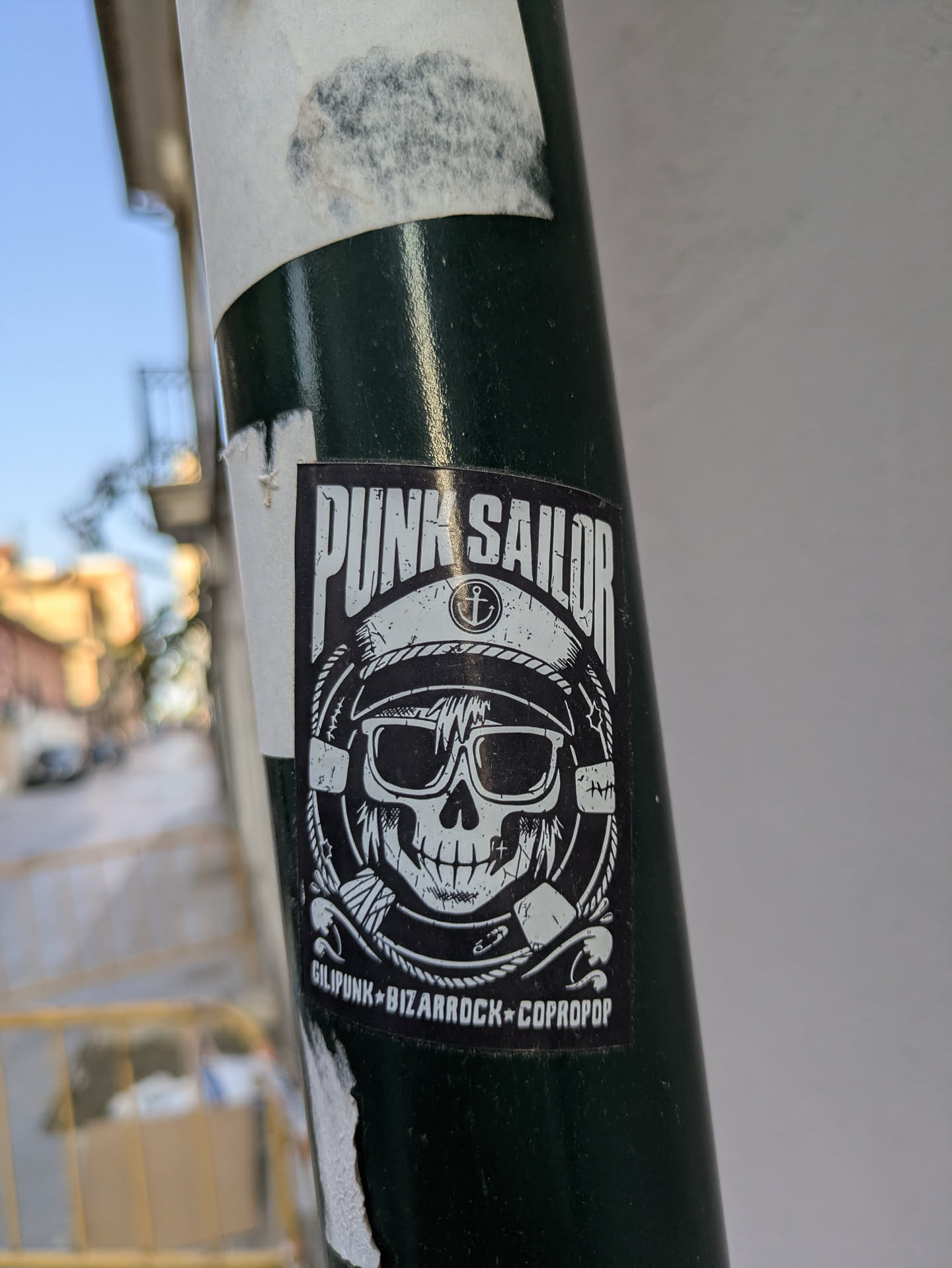 A black and white sticker on a pole features a skull wearing a sailor hat and sunglasses. The text 'Punk Sailor' is at the top, and 'Gilipunk Bizarrock Copropop' is at the bottom. The skull is designed in a punk style.