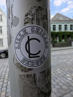 Street sticker The sticker is round with a white background and thick black outlines. The words 'LILLE CHICAGO' are written in a curved arc along the top edge, and 'BERGEN' is similarly curved along the bottom edge, both in a bold, blocky sans-serif font. In the middle of the sticker, there is a prominent black stylized logo, appearing as an 'L' integrated into a 'C'. The sticker is adhered to a light-colored, possibly metallic pole, which shows signs of wear and tear, including some peeling and discoloration from previous adhesives or dirt. The background is slightly blurred, revealing parts of buildings and a cobbled street under a bright sky.