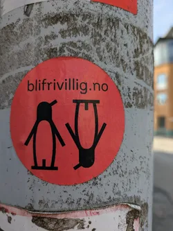 Street sticker A vibrant red, circular sticker is affixed to a textured grey utility pole. The sticker features two stylized, black figures. The figure on the left resembles a simplified, upright penguin. The figure on the right is an inverted, abstract representation of a person, appearing upside down. Above these figures, the black text "blifrivillig.no" is prominently displayed. The sticker's surface has a subtle, granular texture.