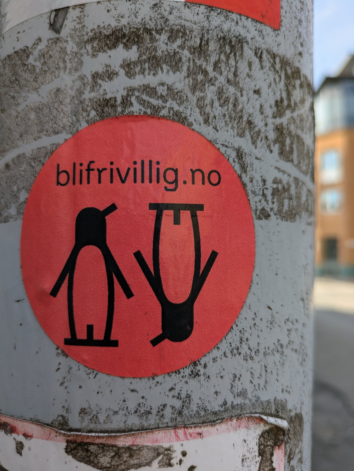 A vibrant red, circular sticker is affixed to a textured grey utility pole. The sticker features two stylized, black figures. The figure on the left resembles a simplified, upright penguin. The figure on the right is an inverted, abstract representation of a person, appearing upside down. Above these figures, the black text "blifrivillig.no" is prominently displayed. The sticker's surface has a subtle, granular texture.