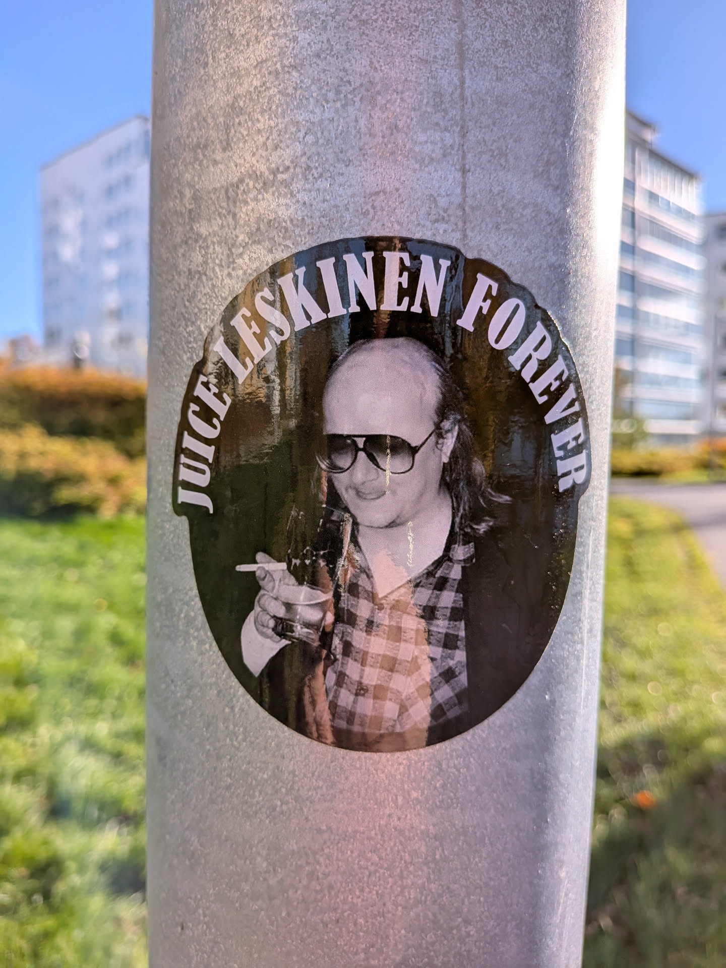 The sticker is circular and features a black background with white text around the perimeter. The text reads 'JUICE LESKINEN FOREVER' in a stylized font. In the center of the sticker is a black and white photograph of a man wearing large, dark sunglasses and a plaid shirt. He is holding a cigarette in his right hand and a glass in his left. The man has a receding hairline and longish hair. The overall aesthetic is reminiscent of a vintage album cover or promotional material.
