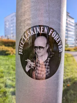 Street sticker The sticker is circular and features a black background with white text around the perimeter. The text reads 'JUICE LESKINEN FOREVER' in a stylized font. In the center of the sticker is a black and white photograph of a man wearing large, dark sunglasses and a plaid shirt. He is holding a cigarette in his right hand and a glass in his left. The man has a receding hairline and longish hair. The overall aesthetic is reminiscent of a vintage album cover or promotional material.