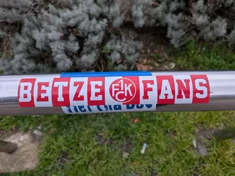 Street sticker A rectangular sticker prominently featuring the text 'BETZE FANS' in bold, white block letters against a vibrant red background. In the center, replacing the space between 'BETZE' and 'FANS', is a circular logo with a white background and a red outline, containing the stylized red letters '1. FCK'. The sticker is applied horizontally on a silver metal railing, with a small strip of blue tape visible directly above it. A partially obscured blue and white sticker is visible underneath the main sticker.