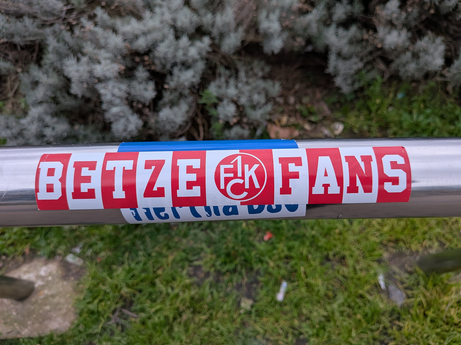A rectangular sticker prominently featuring the text 'BETZE FANS' in bold, white block letters against a vibrant red background. In the center, replacing the space between 'BETZE' and 'FANS', is a circular logo with a white background and a red outline, containing the stylized red letters '1. FCK'. The sticker is applied horizontally on a silver metal railing, with a small strip of blue tape visible directly above it. A partially obscured blue and white sticker is visible underneath the main sticker.