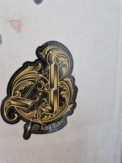 Street sticker A gold and black sticker with a stylized logo featuring the letters "A" and "I" intertwined within ornate, swirling flourishes.  The sticker includes the Instagram handle @Azek_Tattoo at the bottom.