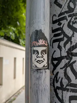 Street sticker A rectangular black and white sticker depicting the stylized, high-contrast face of Andre the Giant. It is pasted on a grey metal pipe with hand-written graffiti above it.
