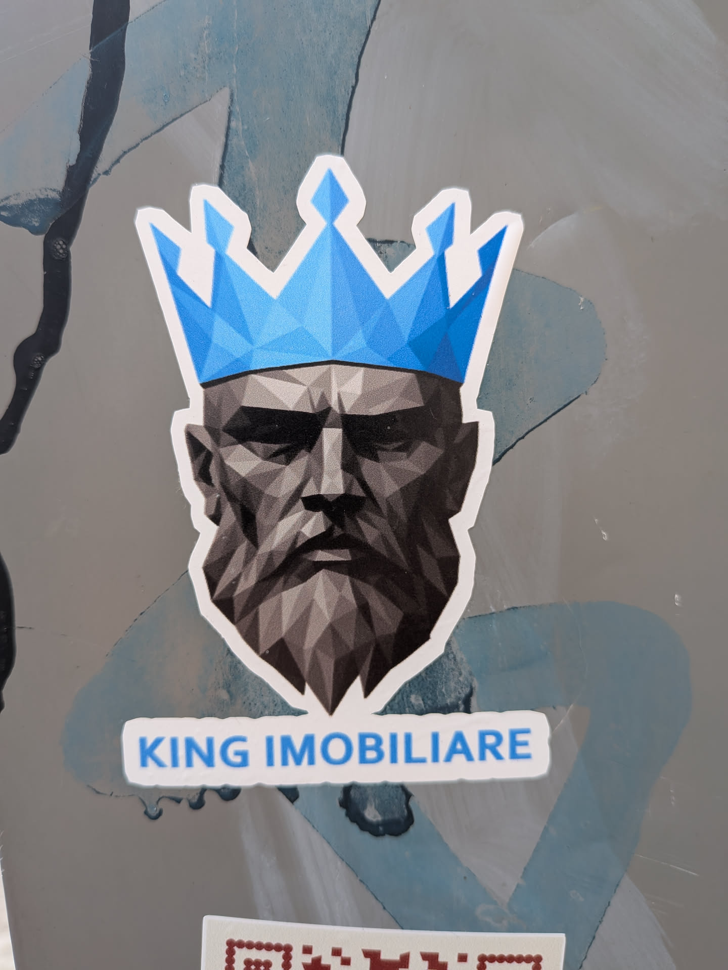 A die-cut sticker featuring a grey, low-poly illustration of a bearded man wearing a bright blue polygonal crown. Below the portrait, the text 'KING IMOBILIARE' is printed in blue capital letters.