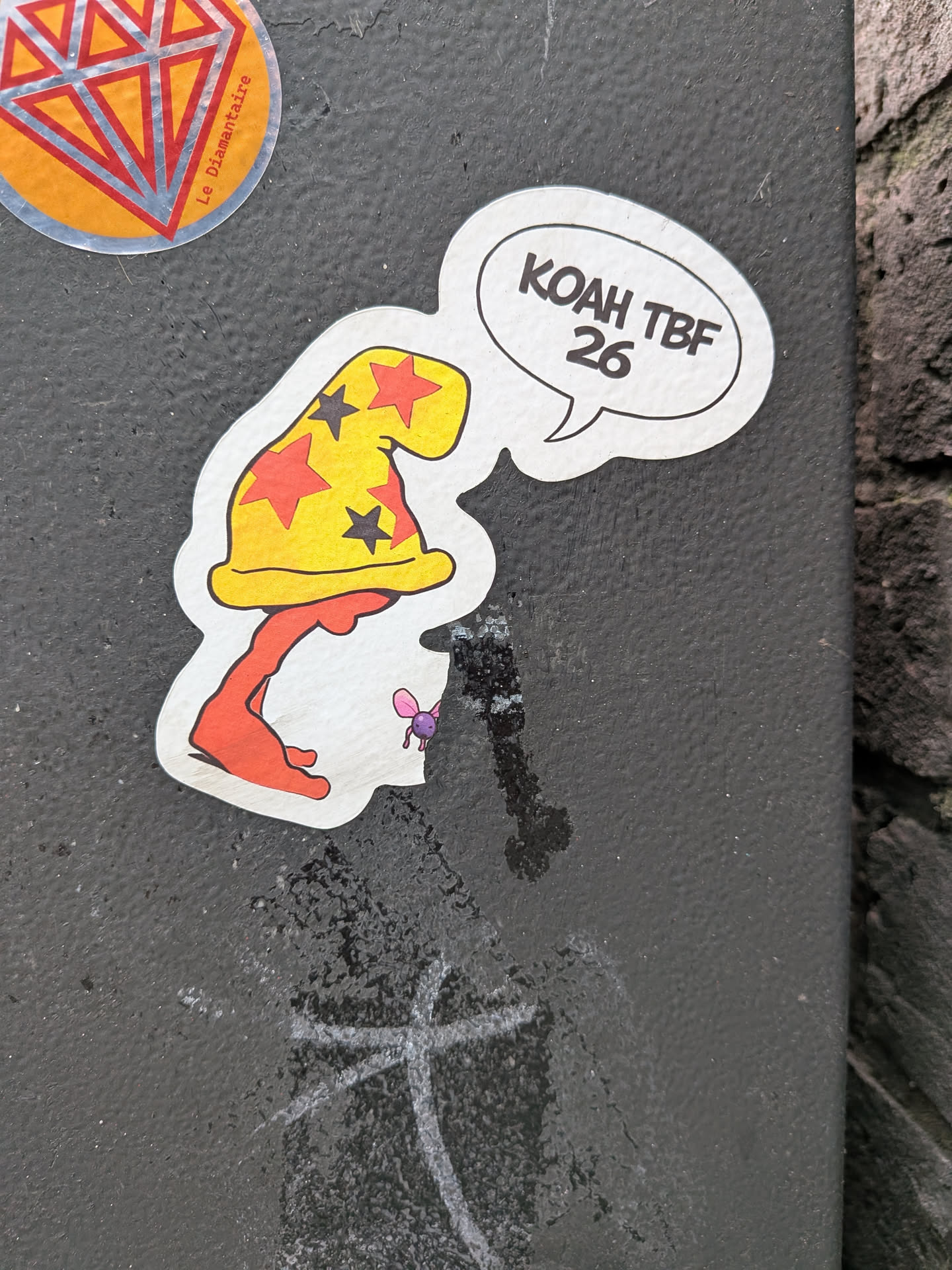 The sticker features a whimsical, cartoon-style character with a prominent yellow wizard hat adorned with red and black stars. The character's body is an elongated, orange, somewhat amorphous shape, possibly resembling legs or a base. A white speech bubble with a black outline emanates from the character's right side. A small, purple, winged insect-like creature is positioned near the base of the orange body. The sticker has a distinct white border around its irregular, die-cut shape.