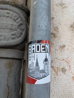 Street sticker The sticker shows a drawing of the city of Baden, Switzerland, with a black, white and red border. The design includes the city's landmarks such as church spires and clock towers, set against a shield-like background.