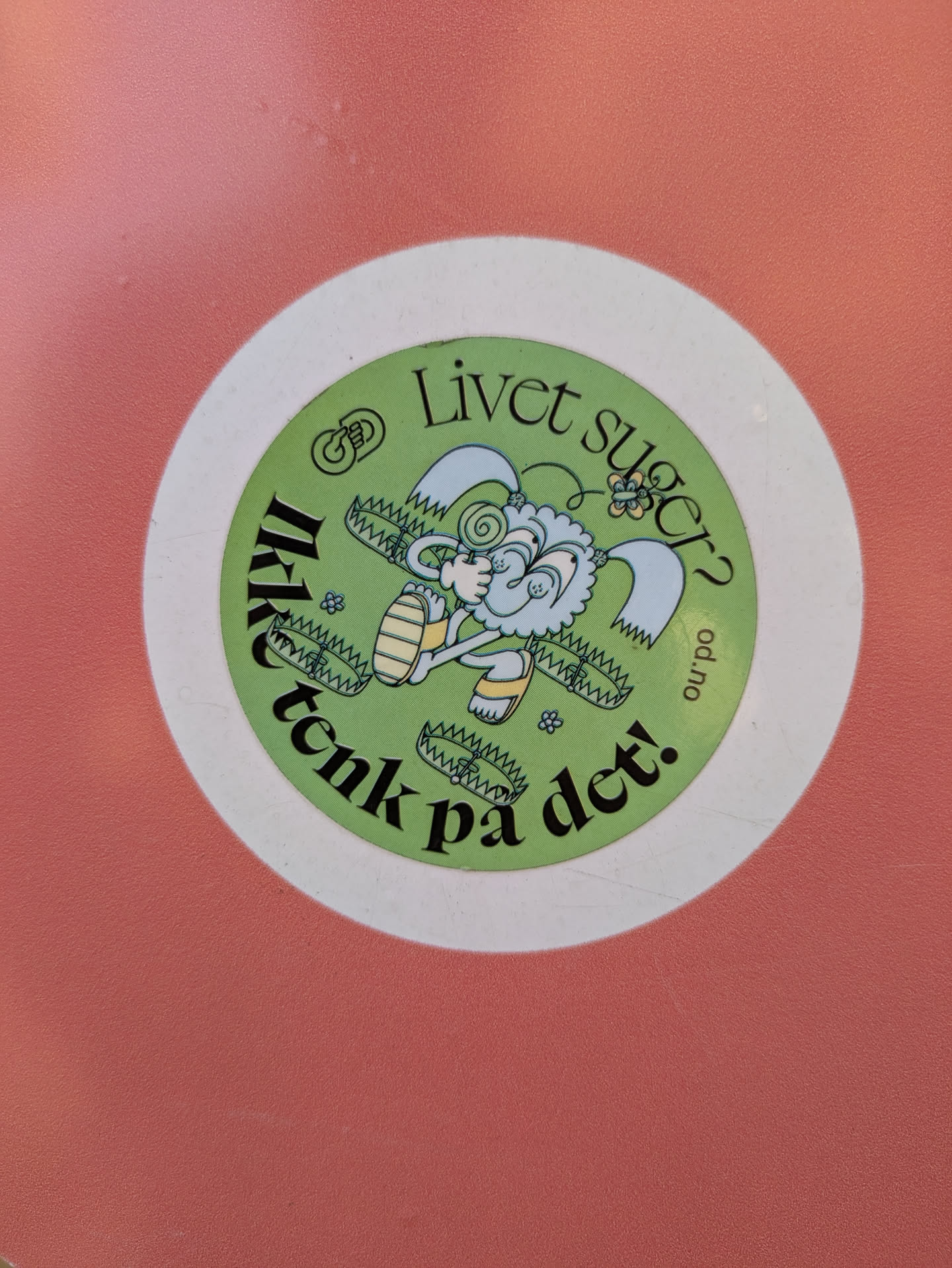 This is a circular sticker with a white outer border and a light green background. The central illustration features a whimsical, fluffy creature, possibly a sheep or a cloud-like being, with light blue and white fur, wearing yellow sandals, and holding a red and white swirl lollipop. Several open bear traps are depicted around the creature's feet. The text "Livet suger?" (Life sucks?) is curved along the top, and "Ikke tenk på det!" (Don't think about it!) is curved along the bottom, both in black lettering. On the right side, oriented vertically, is the web address "od.no". The background includes subtle wavy lines and small floral elements.