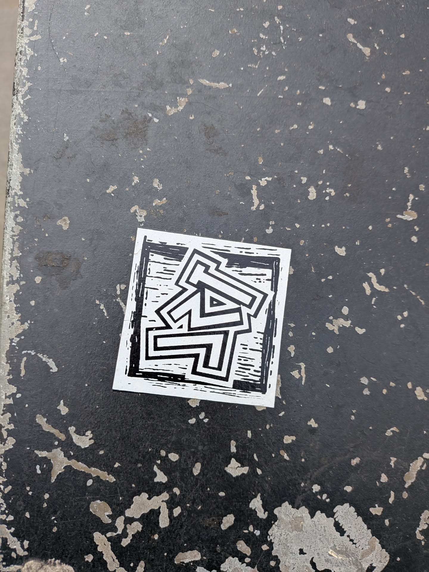 The sticker is a square-shaped, black and white graphic design. It features a central abstract symbol composed of thick, white, outlined geometric shapes that appear to be highly stylized, intertwined letters or numbers (possibly 'TA', 'AT', '47', or '74'). These shapes create a sense of depth with their outlines. The background within the sticker, behind the white shapes, is black with numerous fine white horizontal and vertical lines, giving it a distinct woodcut or linocut print texture. The entire design is framed by a clean white border. The sticker is slightly askew on a dark, heavily weathered surface, possibly metal or concrete, showing significant wear and paint chips.