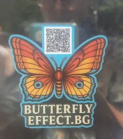 Street sticker The central sticker features a stylized butterfly with wings in shades of orange, yellow, and reddish-brown, outlined by a distinct light blue border. The upper wings are predominantly orange with darker veins, transitioning to yellow and reddish-brown towards the lower parts. The lower wings are mainly yellow with orange and reddish-brown accents, including a small blue circle with a black dot on each. Above the butterfly's body, a black and white QR code is prominently displayed against a light blue background. Below the butterfly, the text "BUTTERFLY EFFECT.BG" is written in bold, light blue capital letters with a dark outline, matching the butterfly's border. The overall design has a clean, somewhat geometric aesthetic.