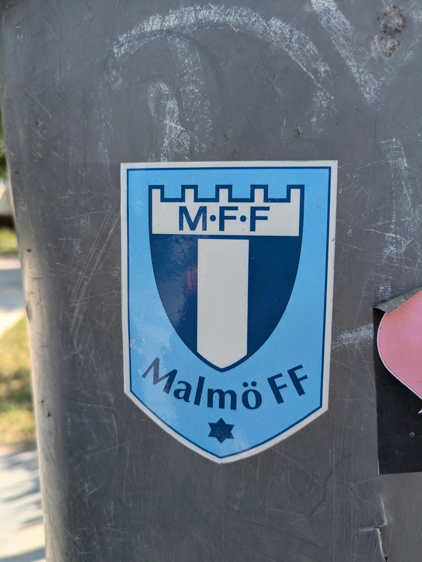 A light blue and dark blue sticker of the Malmö FF football club on a metal pole. The sticker is shaped like a shield. The text reads 'M.F.F.' at the top and 'Malmö FF' at the bottom, with a star underneath.