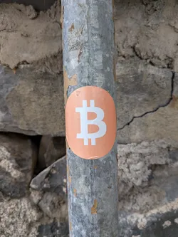Street sticker An oval orange sticker featuring a white Bitcoin symbol, adhered to a weathered metal pole.
