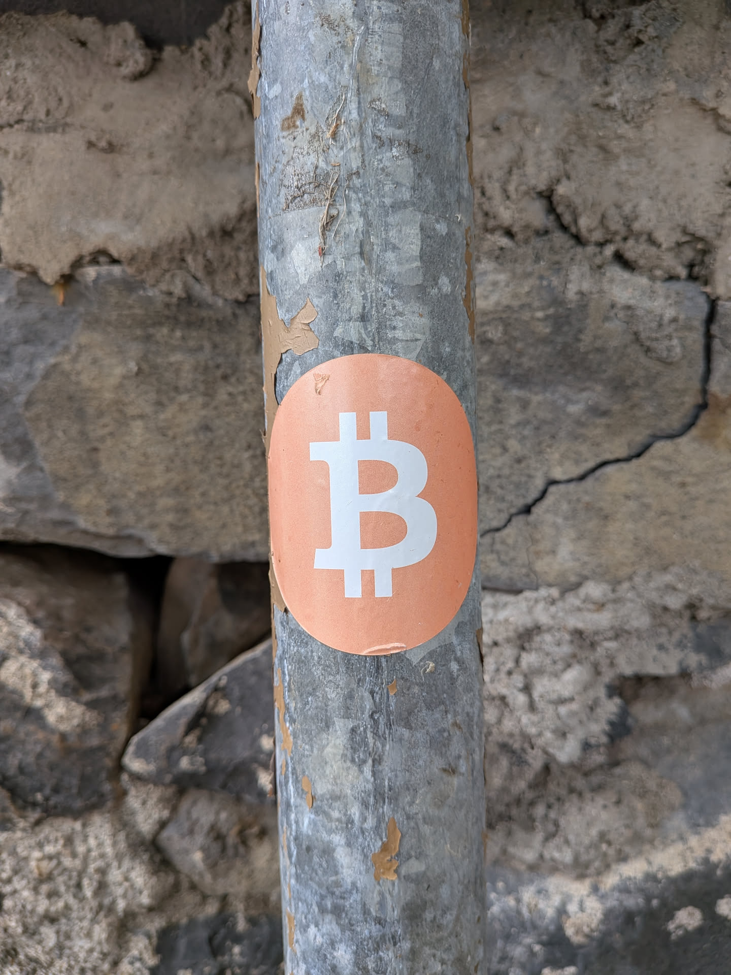 An oval orange sticker featuring a white Bitcoin symbol, adhered to a weathered metal pole.
