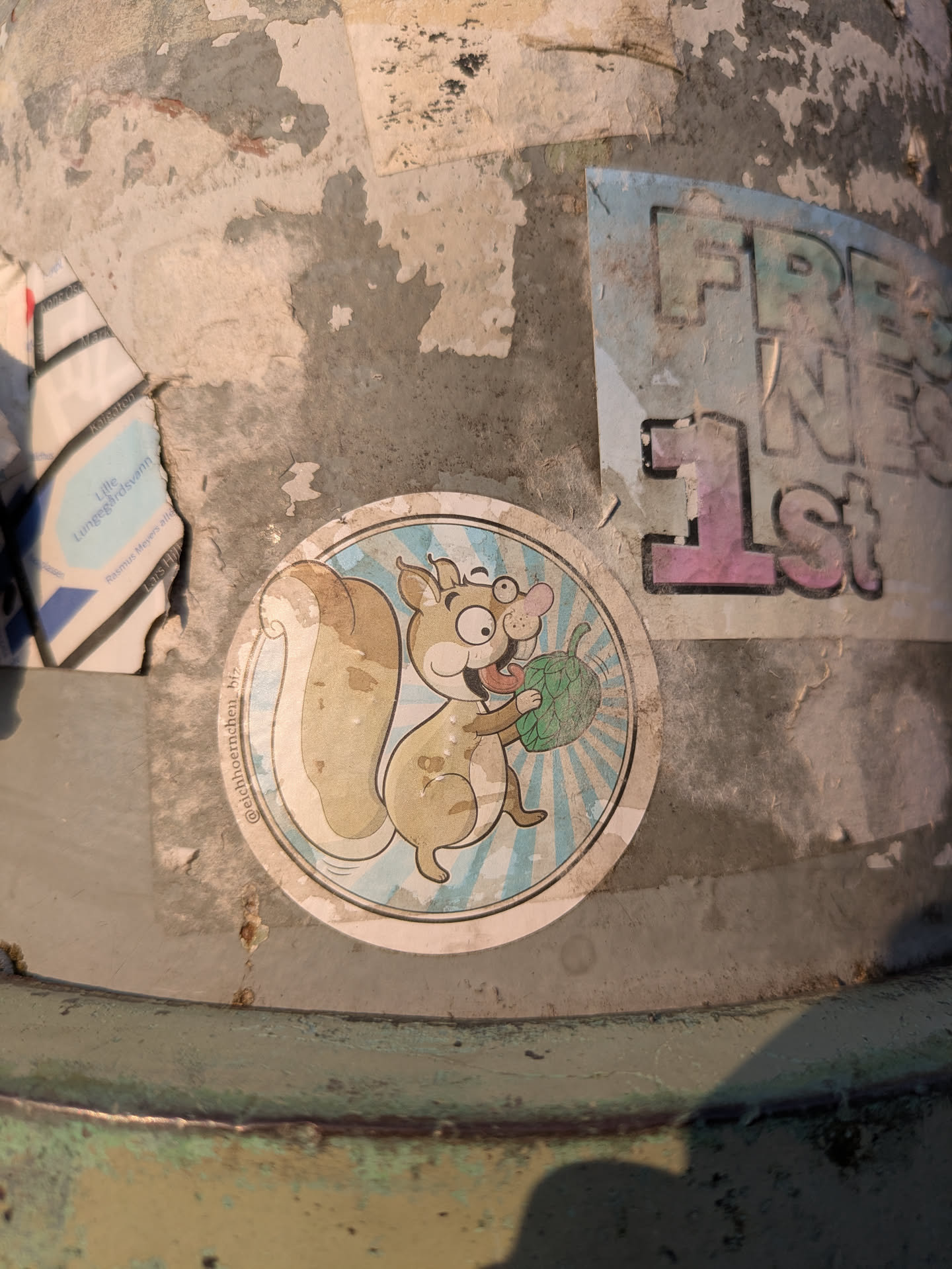 A circular sticker featuring a cartoon squirrel with large, expressive eyes and spectacles. The squirrel, light brown in color, has its tongue playfully sticking out and is holding a green, textured object resembling an acorn or a small fruit. The background of the sticker consists of radiating light blue and white stripes. Along the left edge of the circular design, the text "@eichhoernchen.biz" is printed vertically. The sticker is affixed to a weathered, peeling pole.