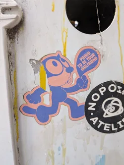 Street sticker A die-cut sticker featuring a Felix the Cat-style character rendered in blue and pink. The character is walking and pointing to a speech bubble containing text. The sticker shows some wear and peeling on its left edge.