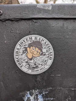 Street sticker A circular sticker featuring a cartoon boy with spiky black hair, light brown skin, and a wide grin, reminiscent of Goku from Dragon Ball. He is wearing a white tank top with a small dark logo and grey pants, raising his right hand to show the middle finger. The background behind the character is a monochrome, distressed illustration of a densely packed urban settlement, possibly a favela. Encircling the top half of the sticker is the text "TORCIDA JOVEM RAPAZIGALO" in dark brown capital letters, and at the bottom, "DESDE 2014" is written in the same style. The sticker has a white/light grey border and appears slightly worn, affixed to a rough, dark grey painted metal surface.