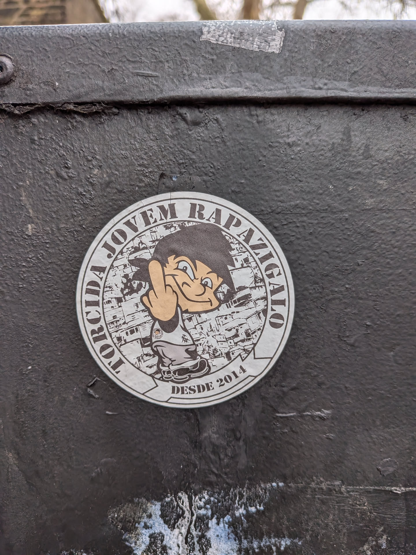 A circular sticker featuring a cartoon boy with spiky black hair, light brown skin, and a wide grin, reminiscent of Goku from Dragon Ball. He is wearing a white tank top with a small dark logo and grey pants, raising his right hand to show the middle finger. The background behind the character is a monochrome, distressed illustration of a densely packed urban settlement, possibly a favela. Encircling the top half of the sticker is the text "TORCIDA JOVEM RAPAZIGALO" in dark brown capital letters, and at the bottom, "DESDE 2014" is written in the same style. The sticker has a white/light grey border and appears slightly worn, affixed to a rough, dark grey painted metal surface.