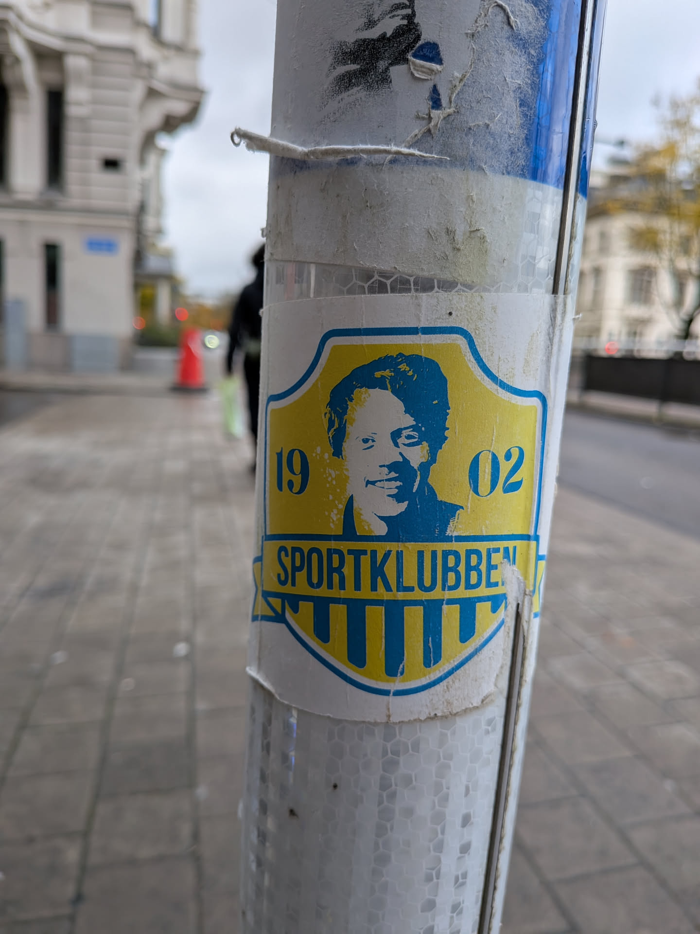 Street sticker The sticker is centrally located on a pole and has a shield-like shape with a blue outline. The background of the shield is yellow. In the upper part of the shield, there is a blue portrait of a person with short, dark hair and a smiling expression. To the left of the portrait is the number '19' in blue, and to the right is the number '02' in blue. Below the portrait, the text 'SPORTKLUBBEN' is written in blue capital letters, with a striped pattern in yellow and blue underneath. The sticker appears to be slightly worn and peeling at the edges.