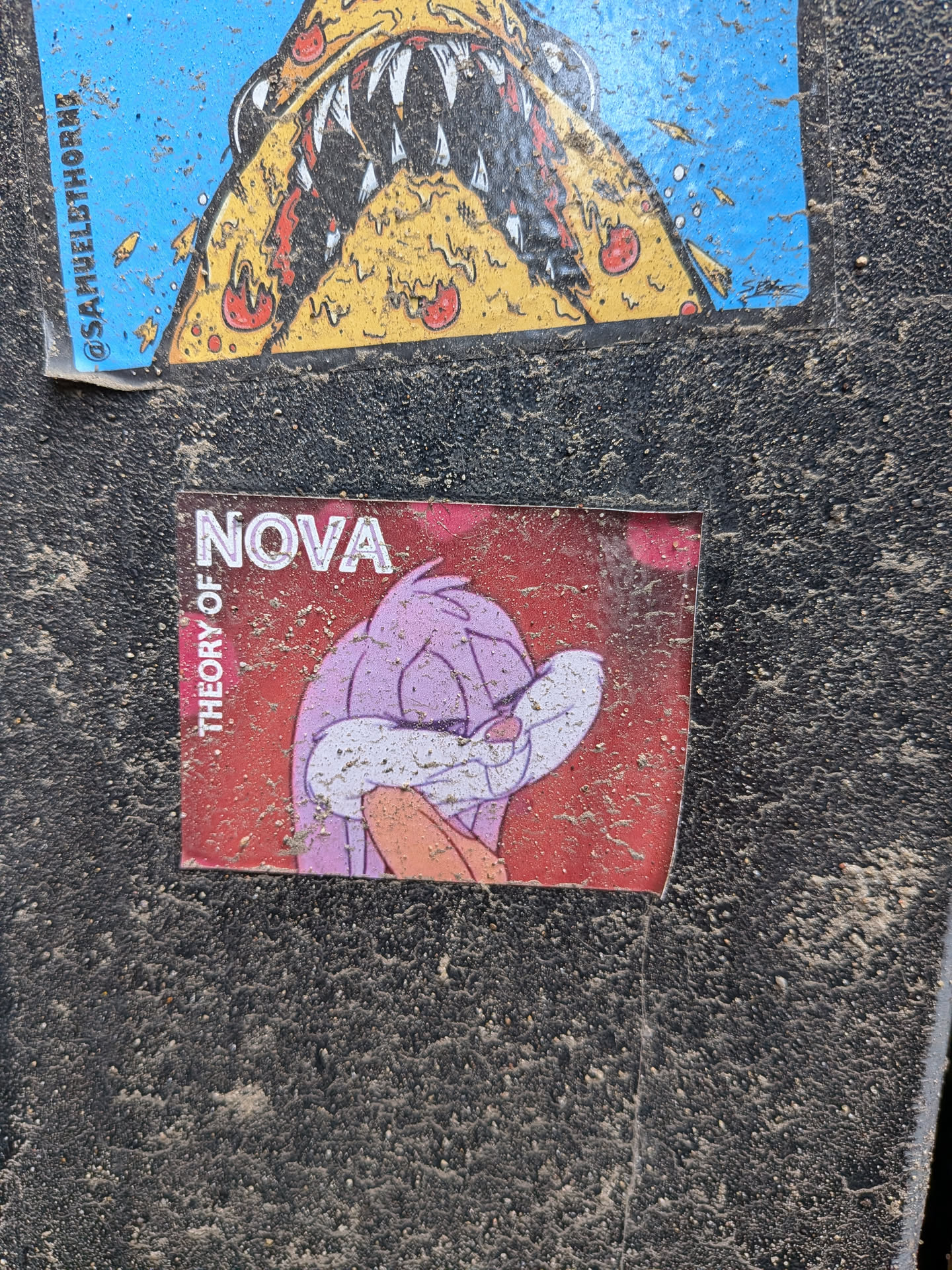 The central sticker features a cartoon character, likely a duck or similar bird, with light purple plumage, a white face, and an orange beak. The character's head is tilted downwards, and a white wing or hand covers part of its face, conveying a pensive or melancholic expression. The background is a faded, somewhat mottled red. White text reads 'NOVA' horizontally at the top right and 'THEORY OF' vertically along the left edge. The rectangular sticker shows signs of wear, dirt accumulation, and slight peeling at the edges, indicating it's affixed to a textured, dark surface.