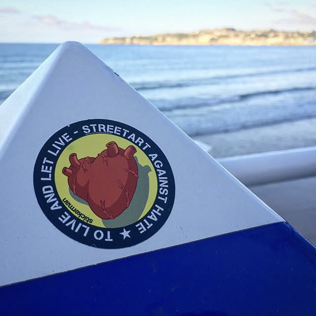 A circular sticker featuring a stylized, reddish-brown anatomical heart at its center, set against a vibrant yellow-green background. The heart is detailed with visible veins and arteries. Encircling the heart is black text on the yellow-green background, which reads 'STREETART AGAINST HATE' at the top and 'TO LIVE AND LET LIVE -' at the bottom. A small, upside-down text 'stickerman' is visible at the very bottom. The entire design is framed by a solid black outer border.