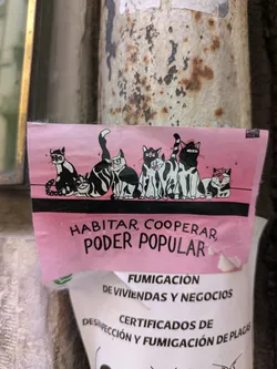 Street sticker The pink sticker shows a row of black and white cartoon cats. Below the cats, the text reads "HABITAR, COOPERAR, PODER POPULAR".  The sticker appears to be on a pole or wall, partially covering another sticker.