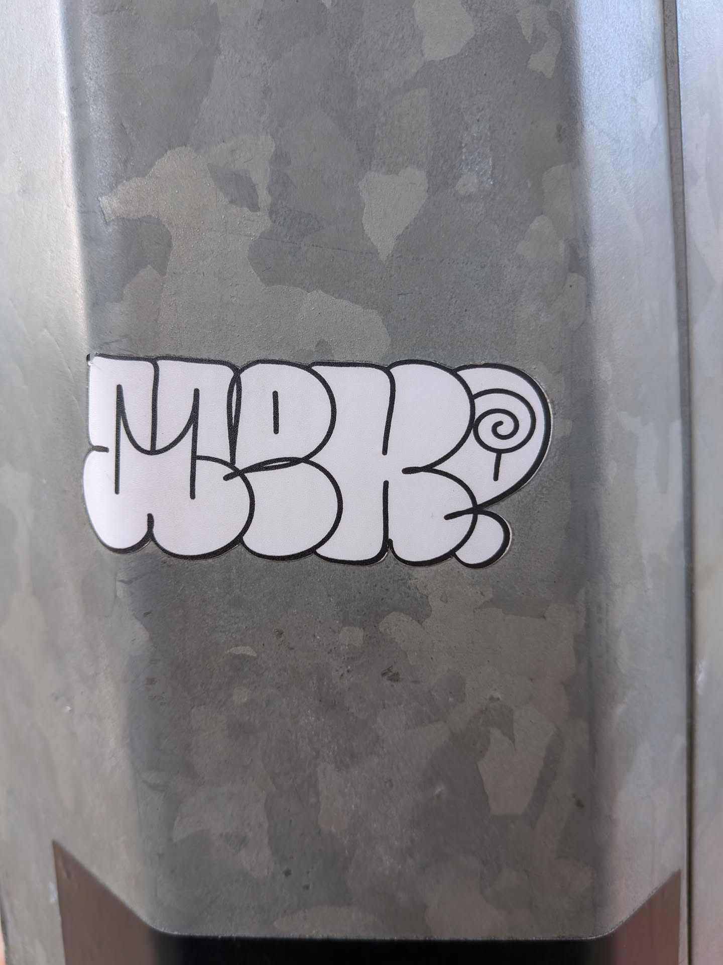 A die-cut vinyl sticker featuring white bubble-style letters with a bold black outline spelling 'MBK' followed by a spiral graphic, placed on a metal surface.