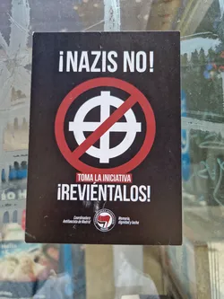Street sticker A sticker with a red circle and a diagonal line crossing out a black symbol (possibly a variation of a Celtic cross). The text in Spanish says: "Nazis No!" ("No Nazis!") and "Toma la iniciativa ¡Reviéntalos!" ("Take the initiative, Smash them!").  The sticker also includes logos for Coordinadora Antifascista de Madrid and Memoria dignidad y lucha.