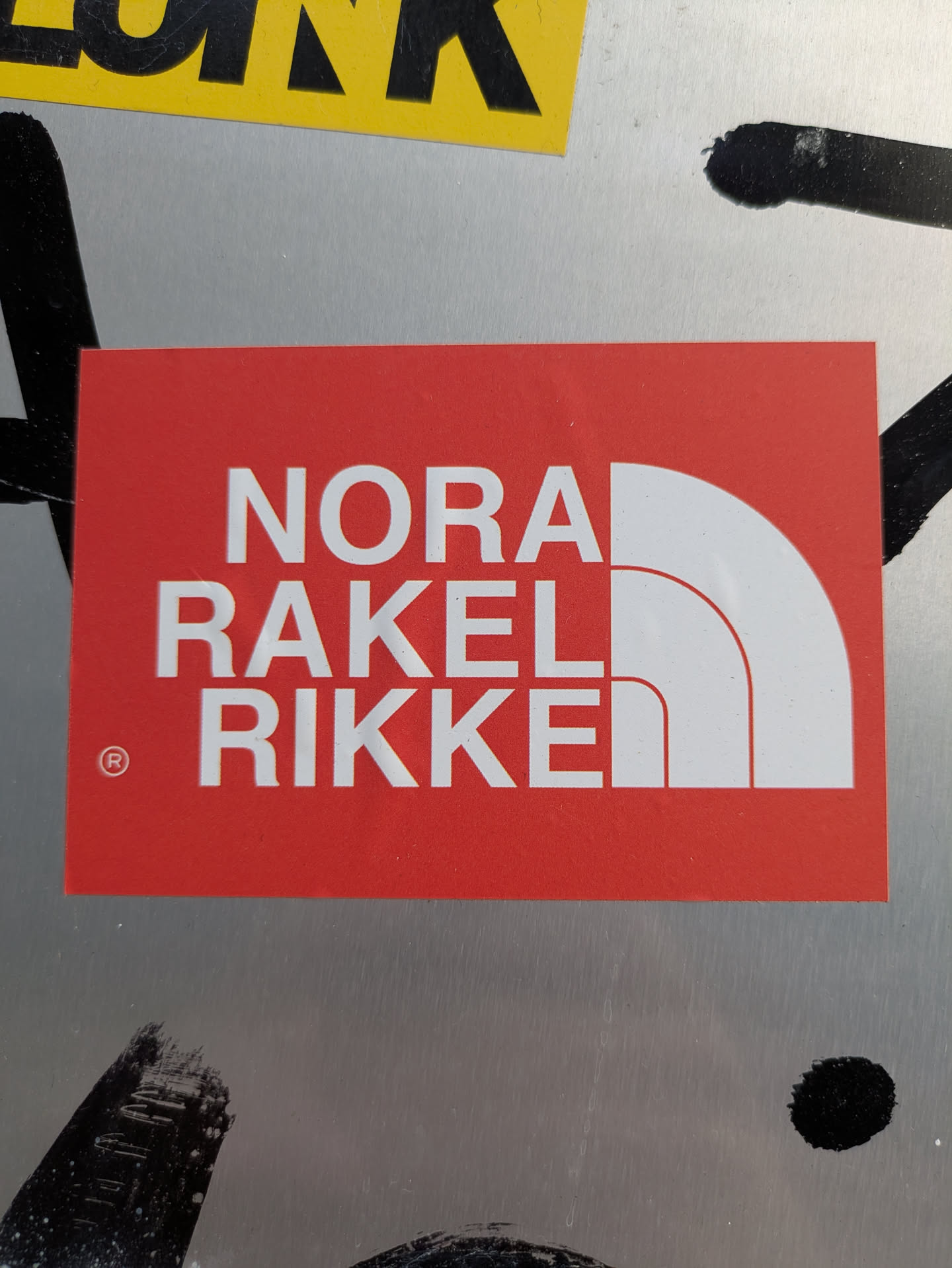This is a red rectangular sticker with white text arranged in three lines: "NORA", "RAKEL", and "RIKKE". The text and the accompanying white abstract semi-circle graphic on the right are a clear parody of The North Face brand logo. A small registered trademark symbol (®) is visible to the left of 'RIKKE'. The sticker is placed on a metallic or silver-colored surface, with some black graffiti marks partially visible around it.