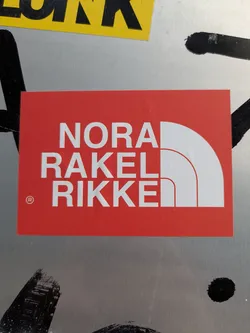 Street sticker This is a red rectangular sticker with white text arranged in three lines: "NORA", "RAKEL", and "RIKKE". The text and the accompanying white abstract semi-circle graphic on the right are a clear parody of The North Face brand logo. A small registered trademark symbol (®) is visible to the left of 'RIKKE'. The sticker is placed on a metallic or silver-colored surface, with some black graffiti marks partially visible around it.