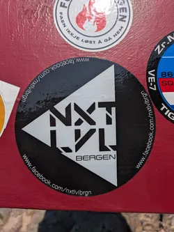 Street sticker This is a black circular sticker with a prominent white logo that reads "NXTLVL" in a stylized, blocky font, positioned within a white triangular design. Below "NXTLVL", the word "BERGEN" is written in a simpler white font. Encircling the edge of the sticker, the Facebook URL "www.facebook.com/nxtlvlbrgn" is printed in white text, indicating a presence on the social media platform. The sticker is applied to a textured, reddish-brown surface.