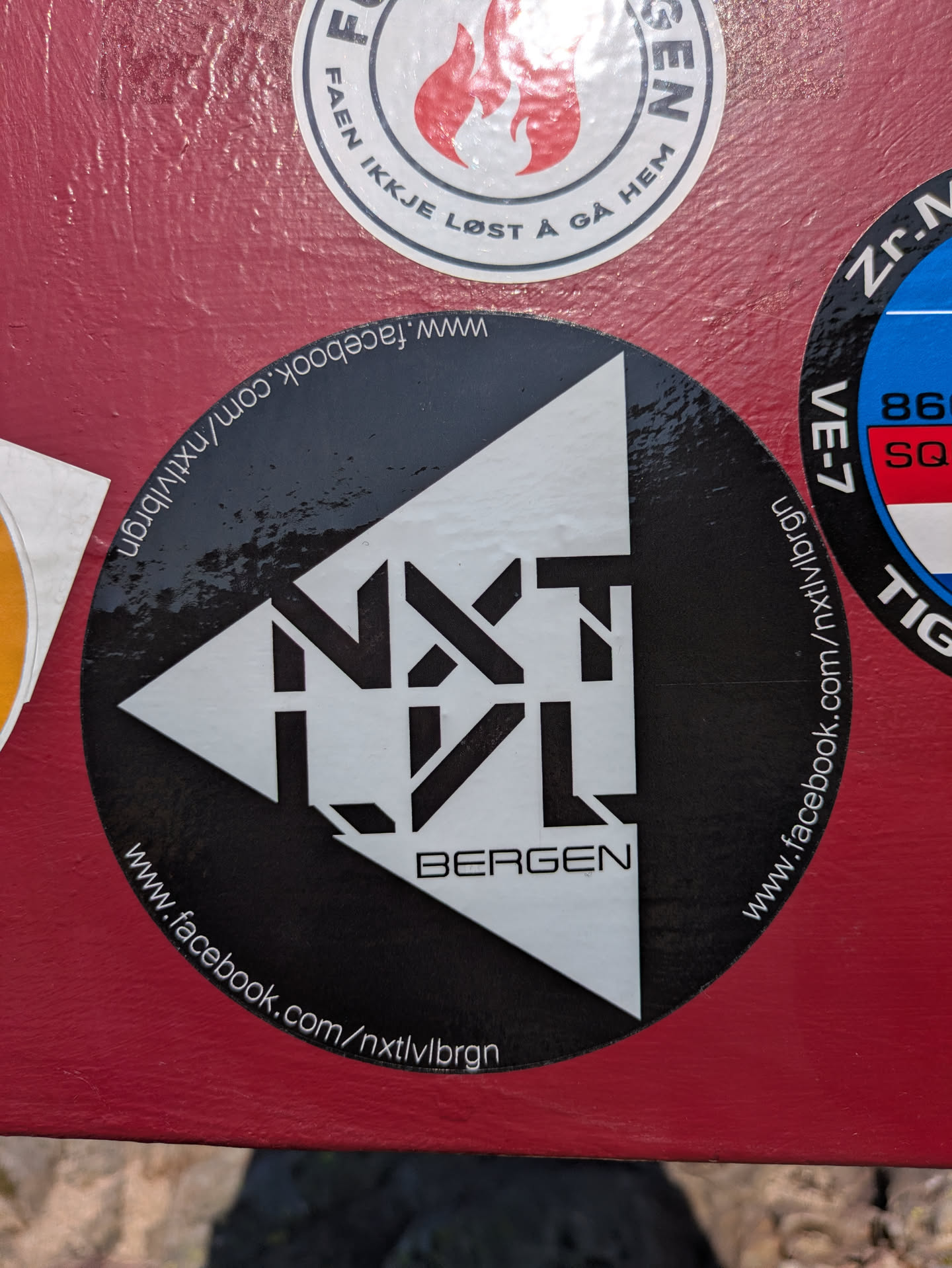 This is a black circular sticker with a prominent white logo that reads "NXTLVL" in a stylized, blocky font, positioned within a white triangular design. Below "NXTLVL", the word "BERGEN" is written in a simpler white font. Encircling the edge of the sticker, the Facebook URL "www.facebook.com/nxtlvlbrgn" is printed in white text, indicating a presence on the social media platform. The sticker is applied to a textured, reddish-brown surface.