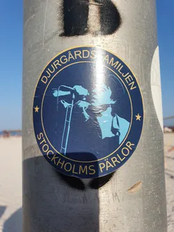 A round sticker featuring a blue silhouette of a person singing into a microphone. The text "DjurgårdsFamiljen" and "Stockholms Pärlör" are written around the image.  The sticker is affixed to a metal pole.